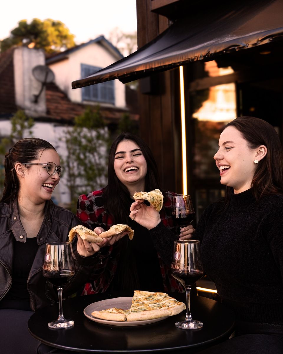 Três amigas riem e comem fatias de pizza com taças de vinho tinto, na área externa