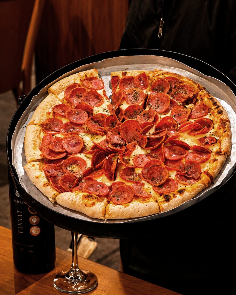 Pizza de calabresa inteira no suporte, com uma garrafa de vinho Reserva Tannat e uma taça na mesa