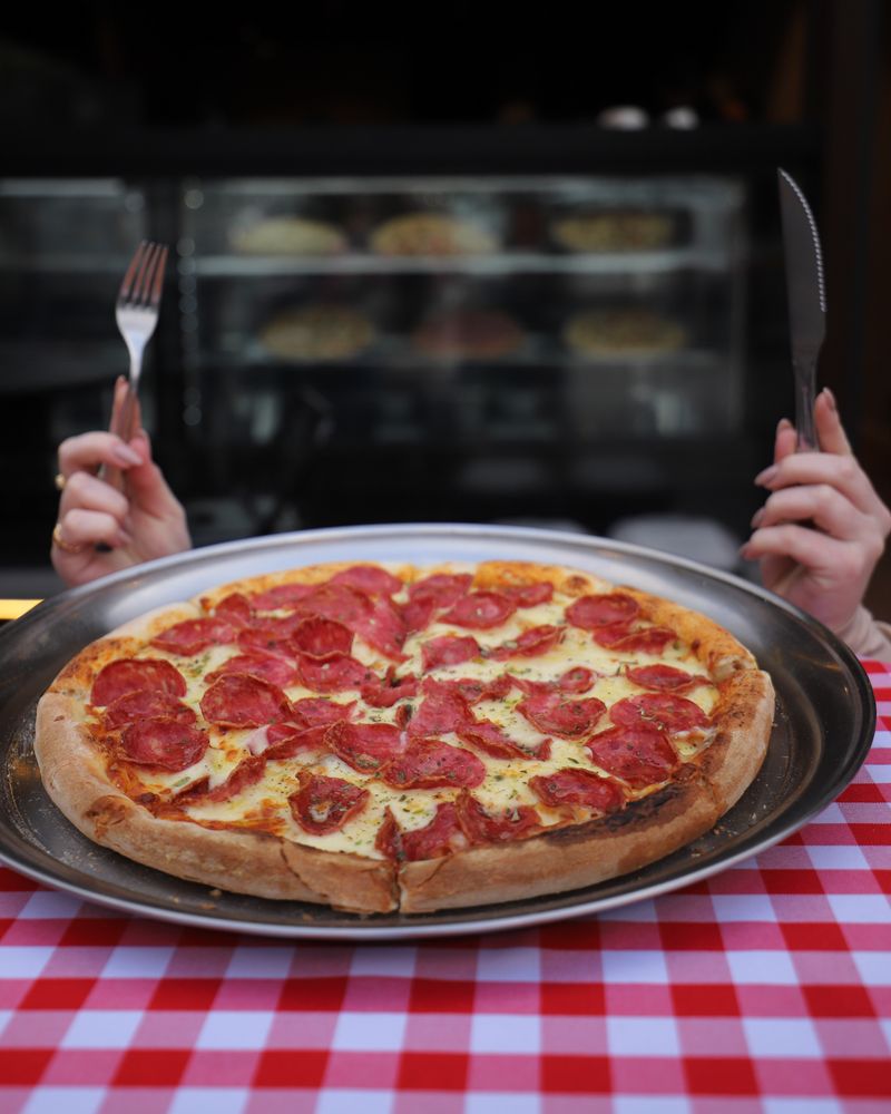Pizza de calabresa inteira na bandeja, sobre a toalha xadrez, com garfo e faca nas mãos