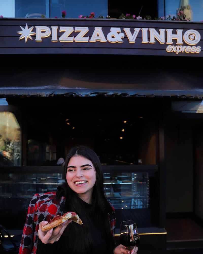 Fachada do Pizza & Vinho Express com o letreiro iluminado e uma cliente com uma fatia de pizza e uma taça de vinho