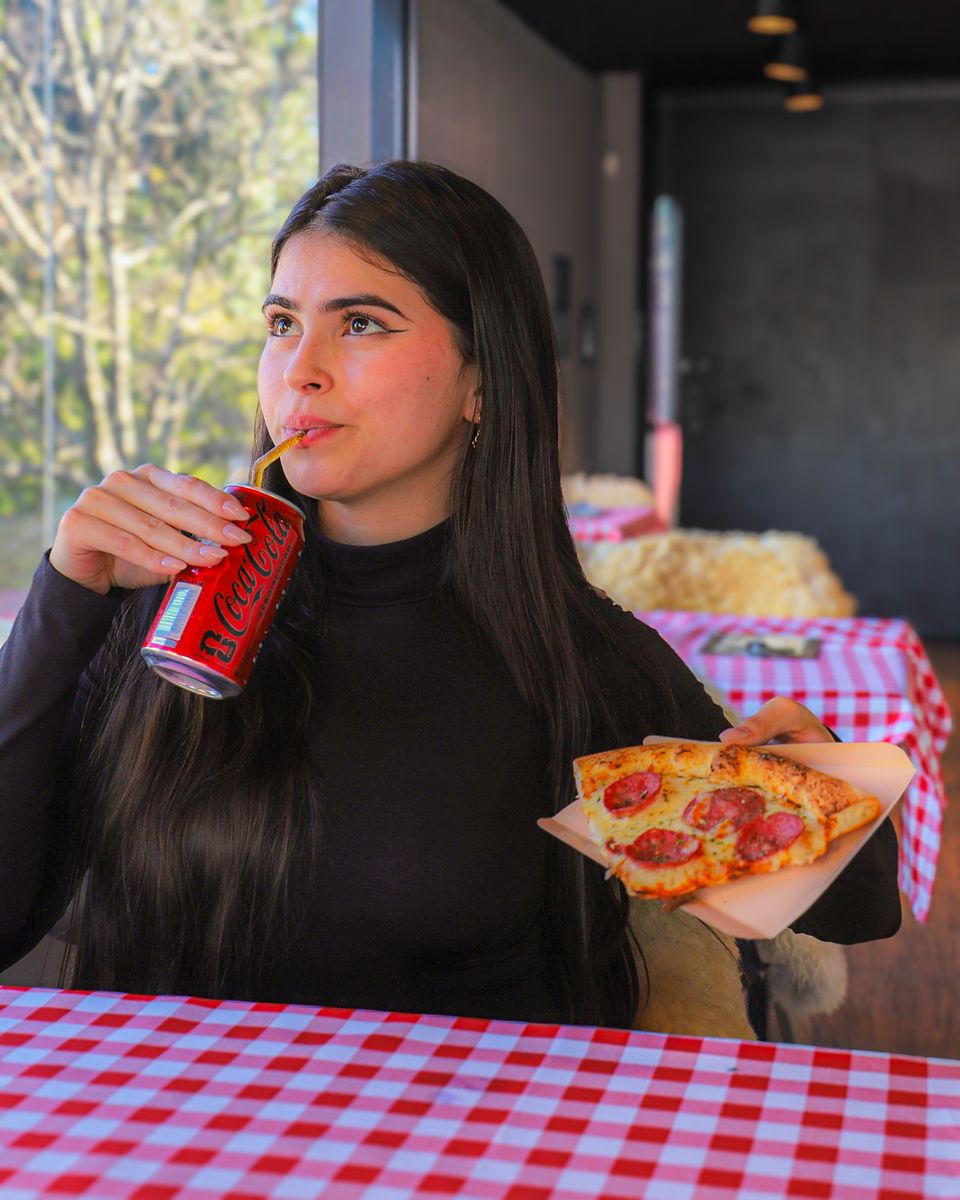 Mulher toma refrigerante de lata com canudo e segura uma fatia de pizza, na mesa com toalha xadrez
